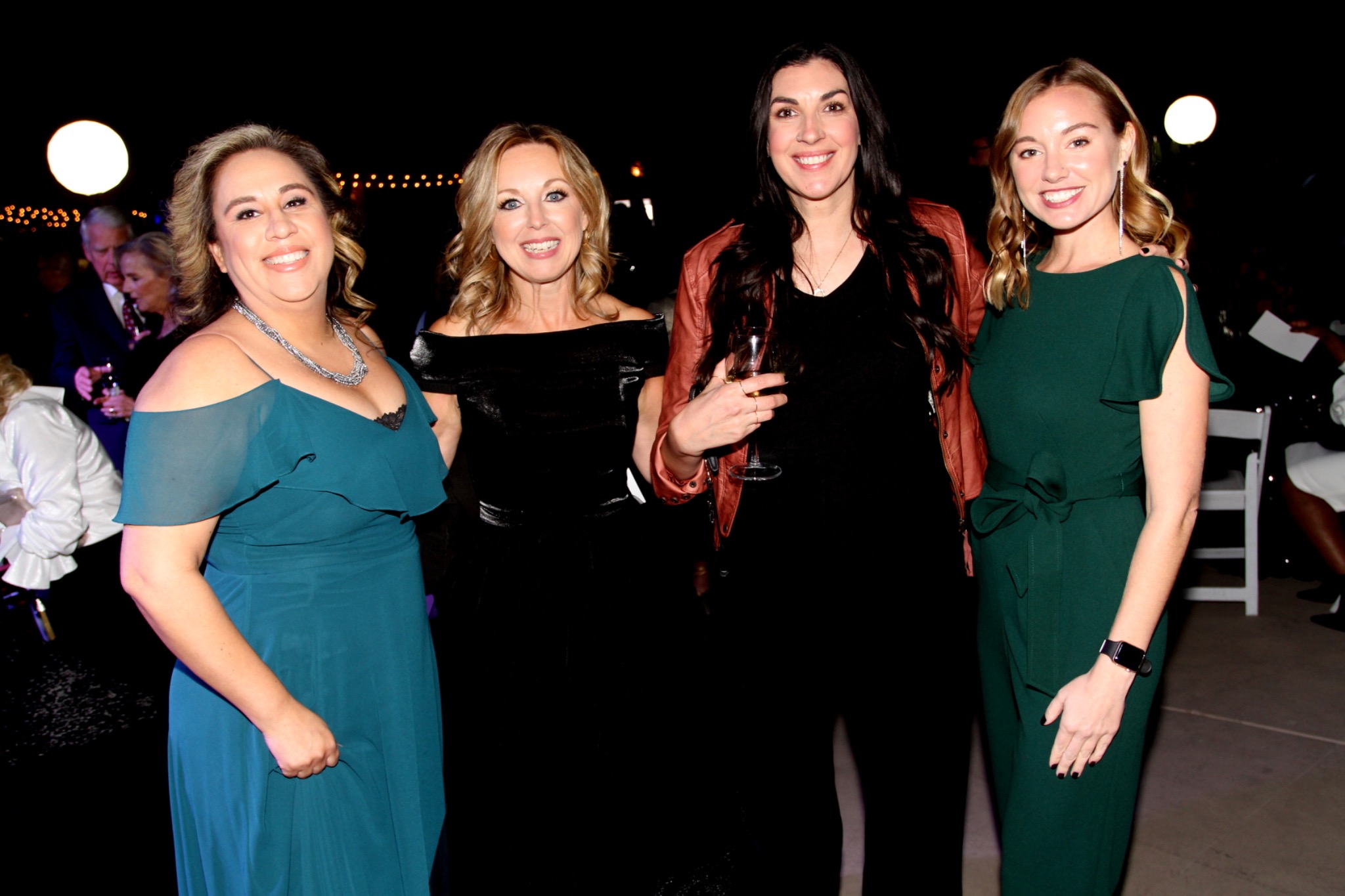 Four guests in teal, green, and black evening dresses gathered outdoors.