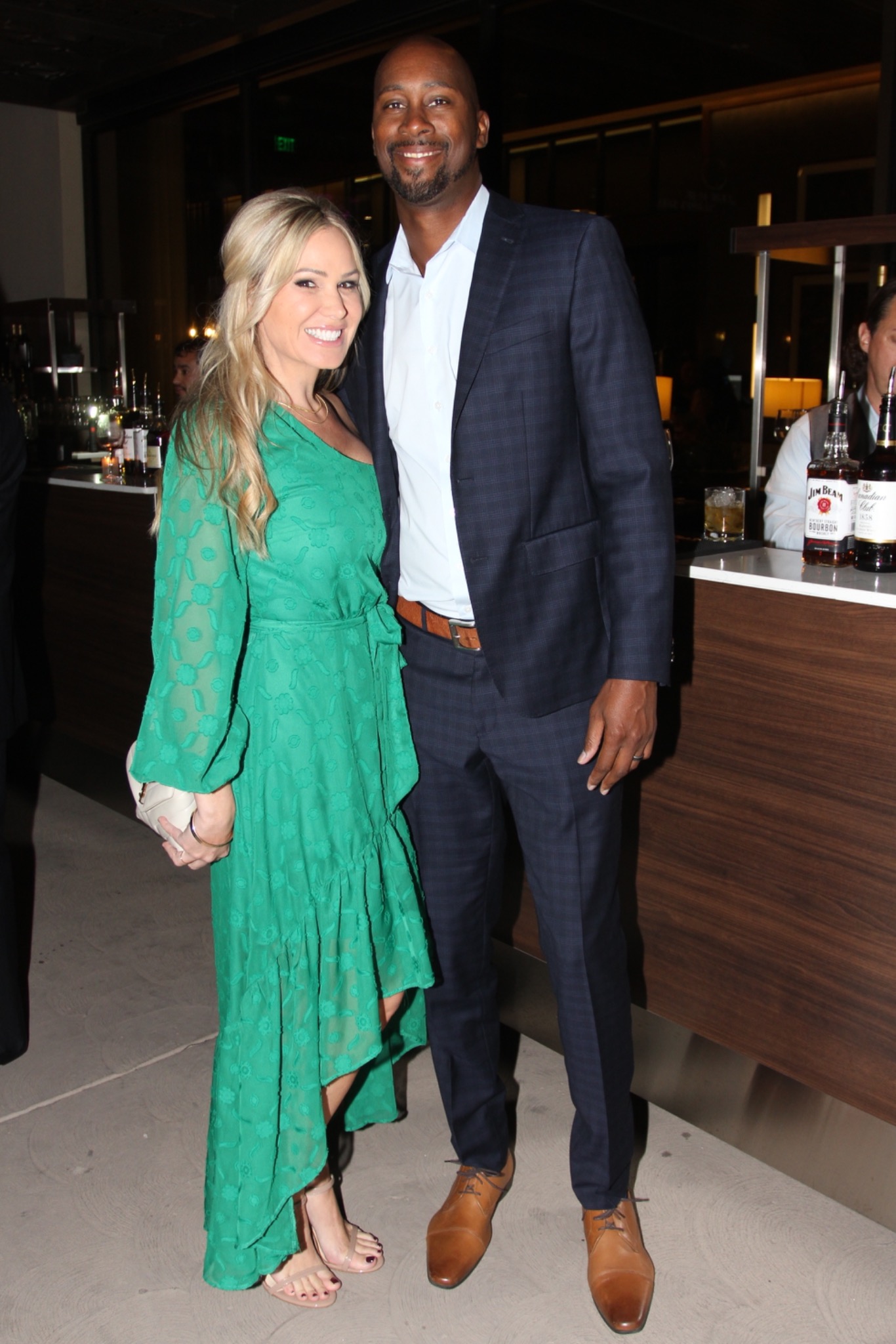 Two guests in a green dress and dark suit standing beside a reception bar.