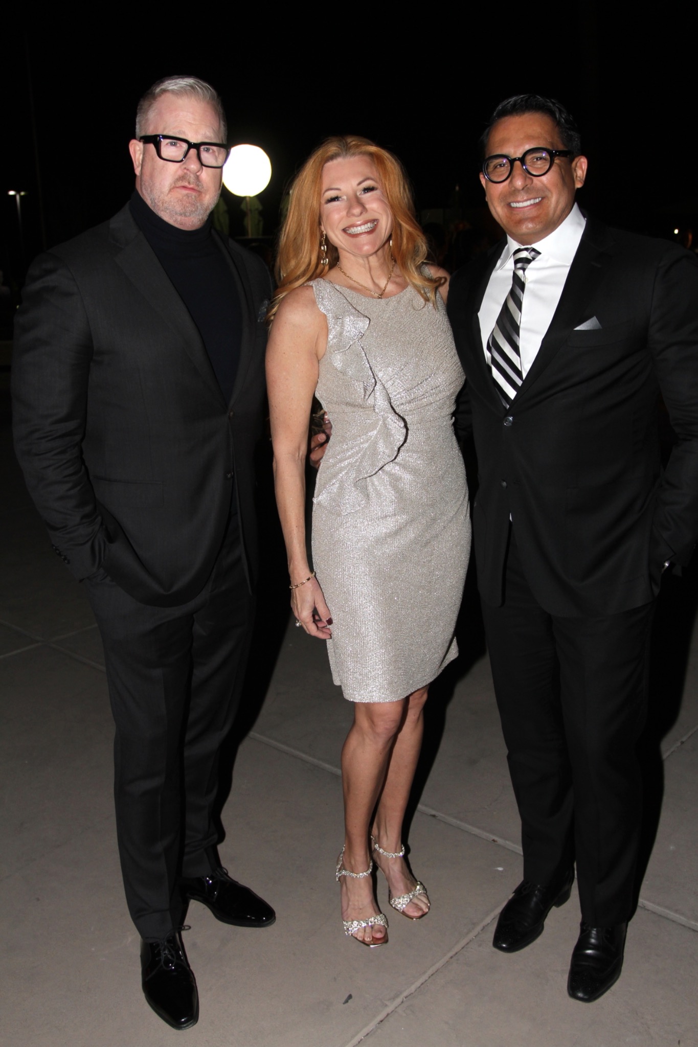 Three guests in dark suits and a silver dress posing outdoors.